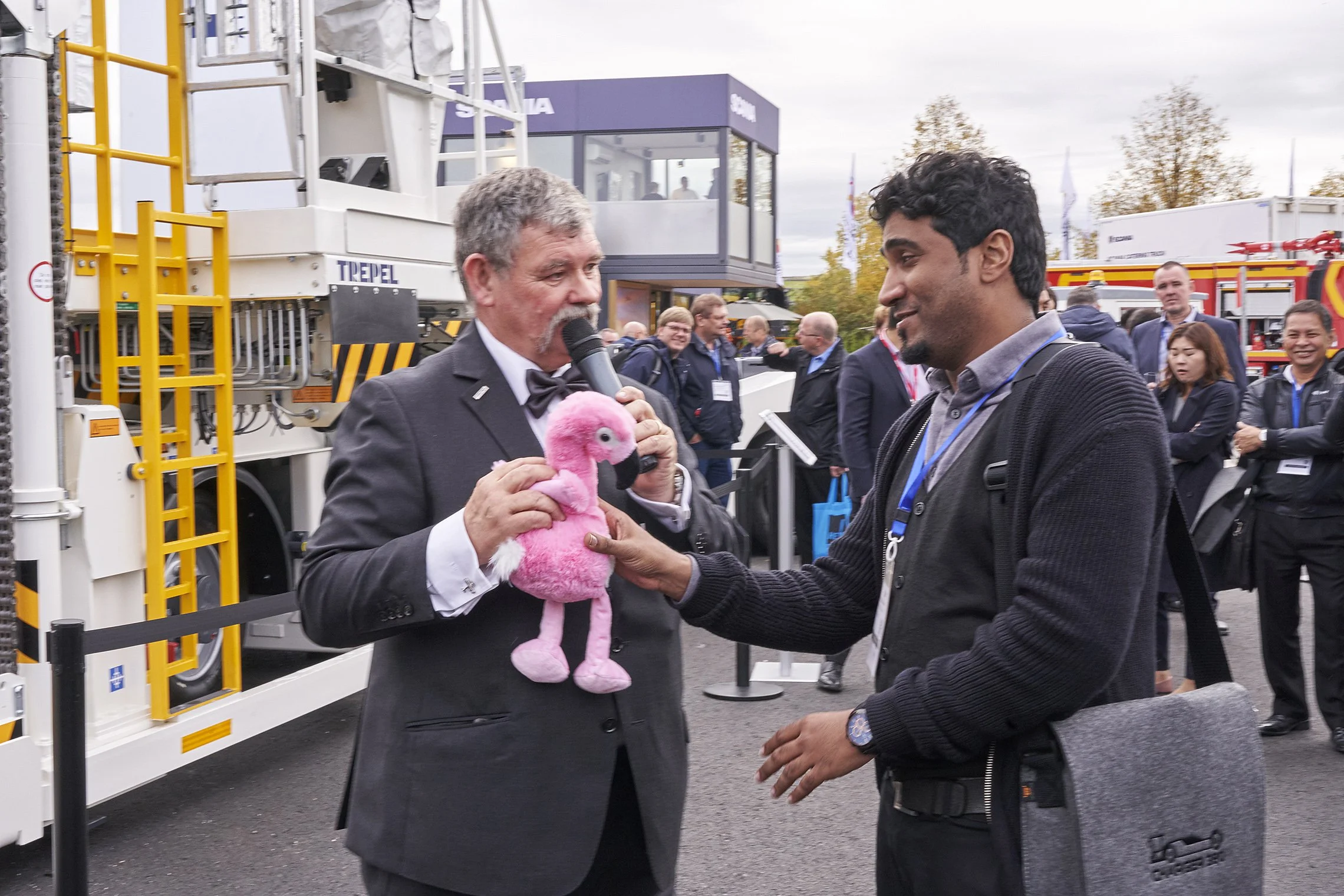 Two men interacting with a pink plush toy during a live presentation in front of a TREPEL high loader