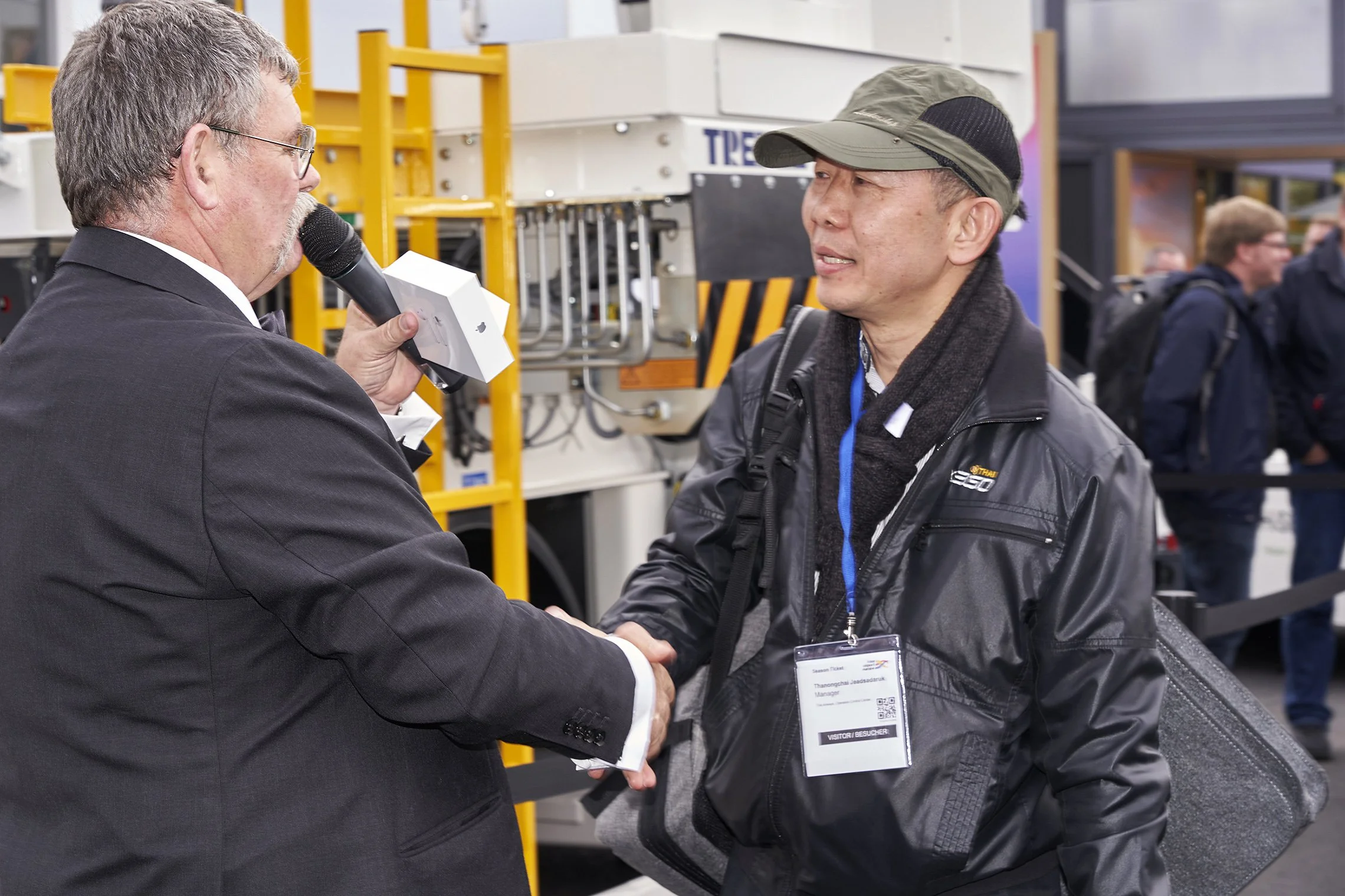 Two men shaking hands, one holding a microphone, in front of a TREPEL high loader