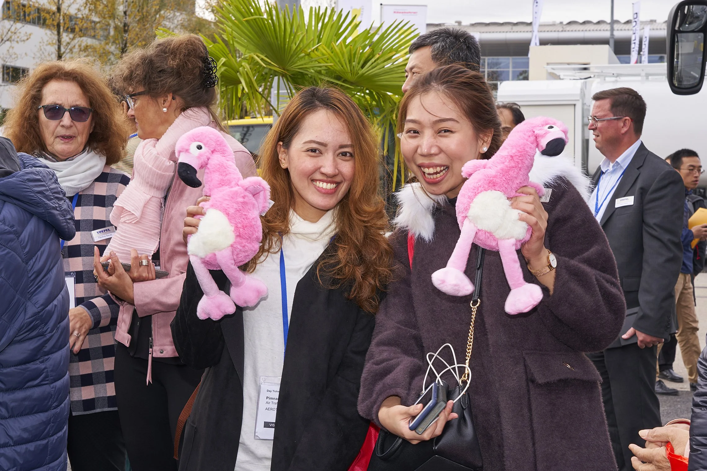 Two women smiling and holding pink flamingo plush toys among other attendees at the TREPEL booth