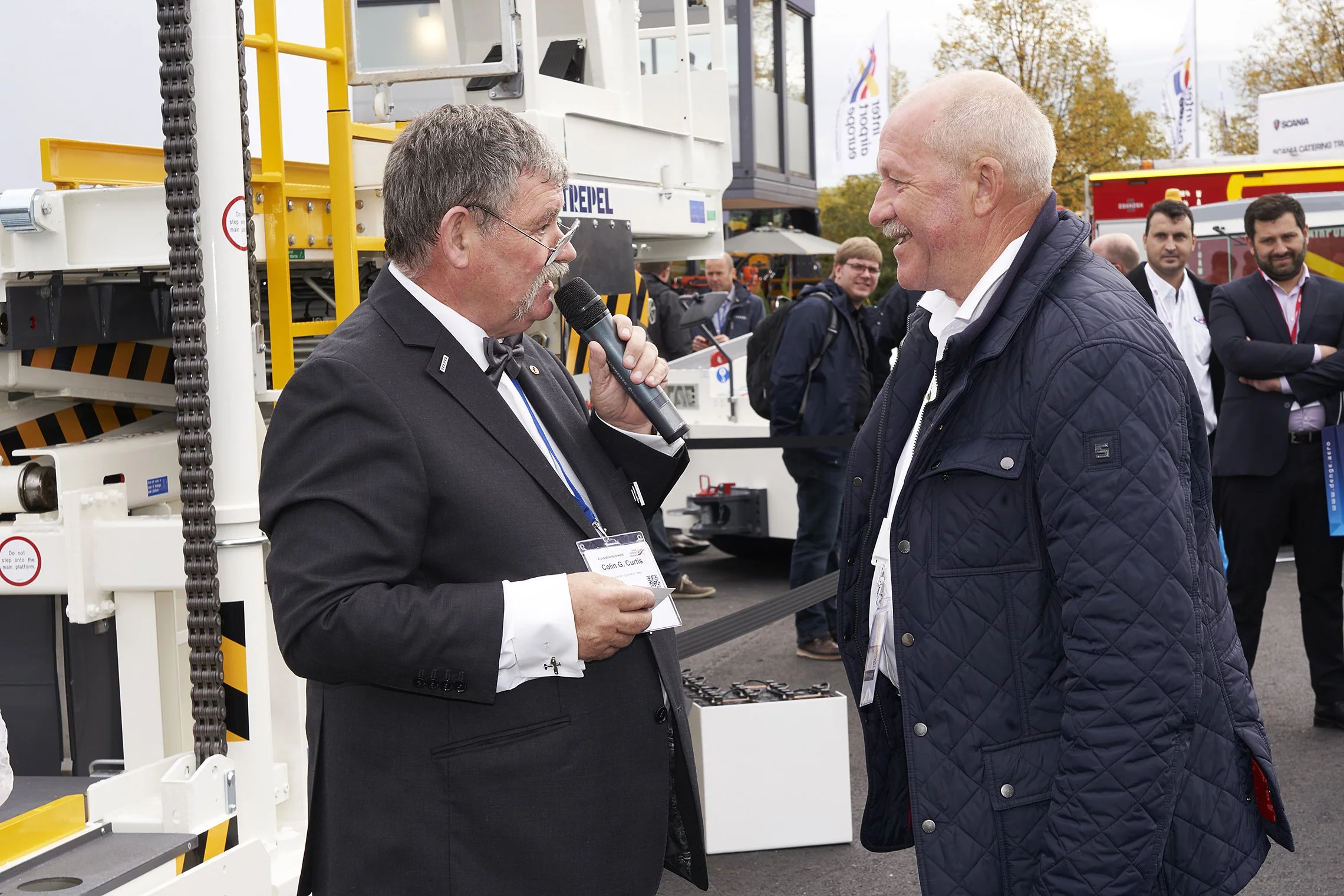 Two men interacting during a live microphone presentation in front of TREPEL ground support equipment