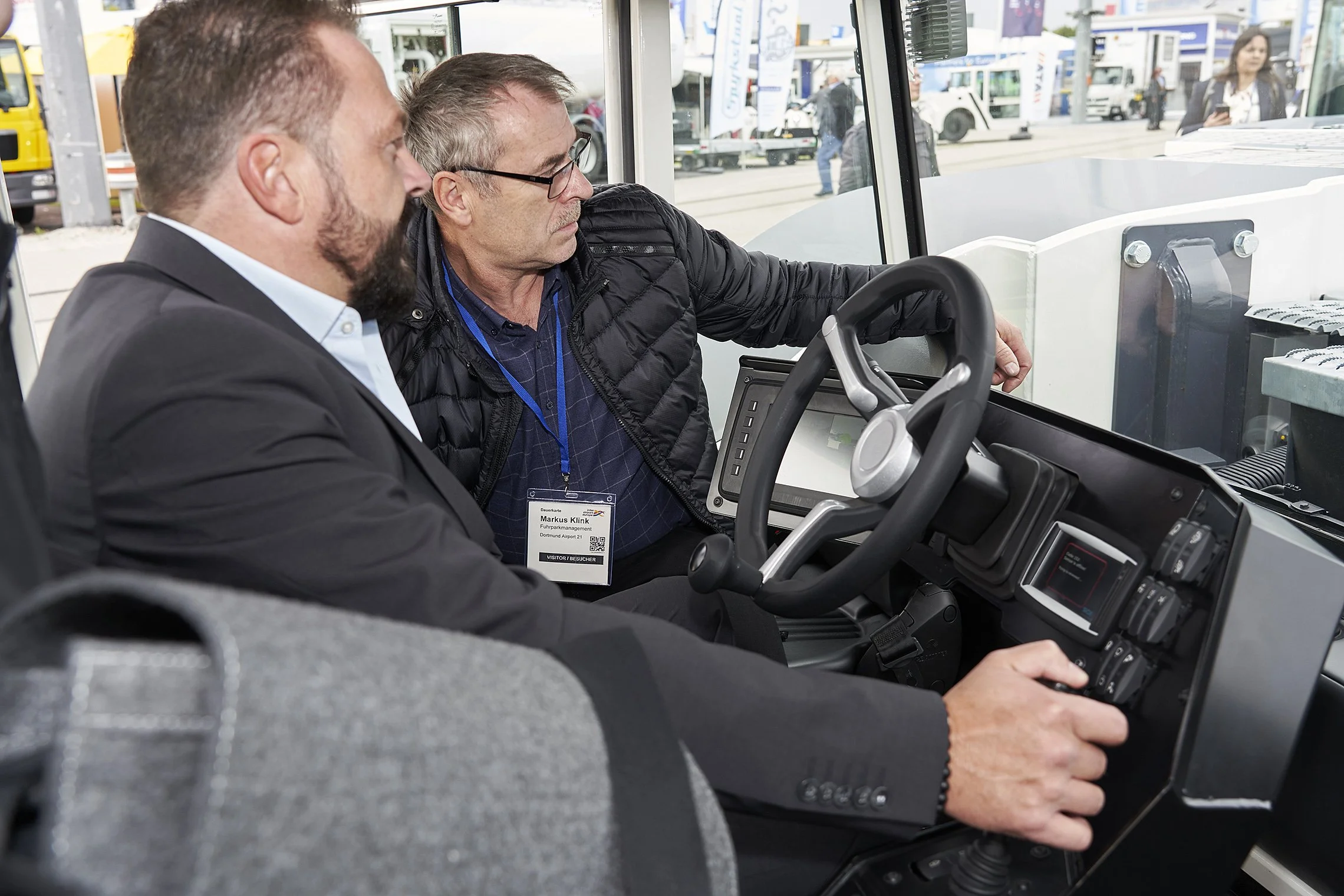 Two men seated inside a TREPEL tow tractor cockpit discussing the vehicle controls
