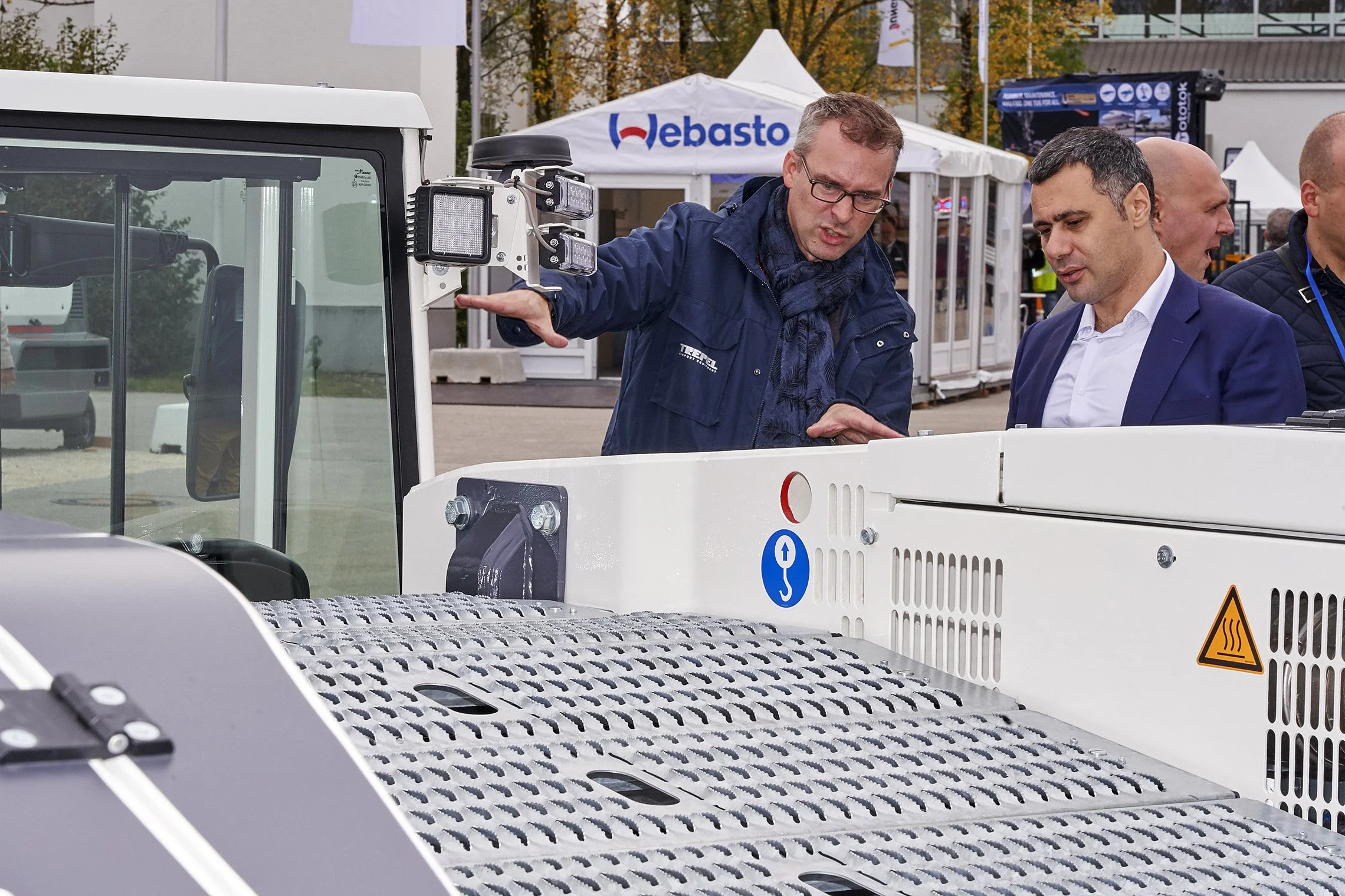 Two men in technical discussion beside a TREPEL aircraft tractor at an outdoor trade fair booth