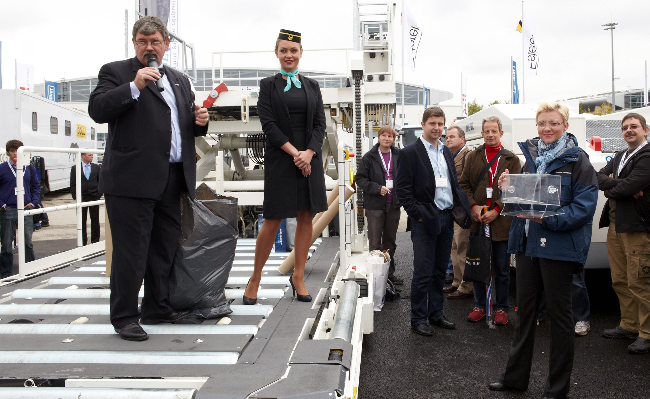 Mann mit Mikrofon und Flugbegleiterin stehen auf einer GSE-Anlage, während eine Frau eine Lostrommel hält und umstehende Besucher zuschauen