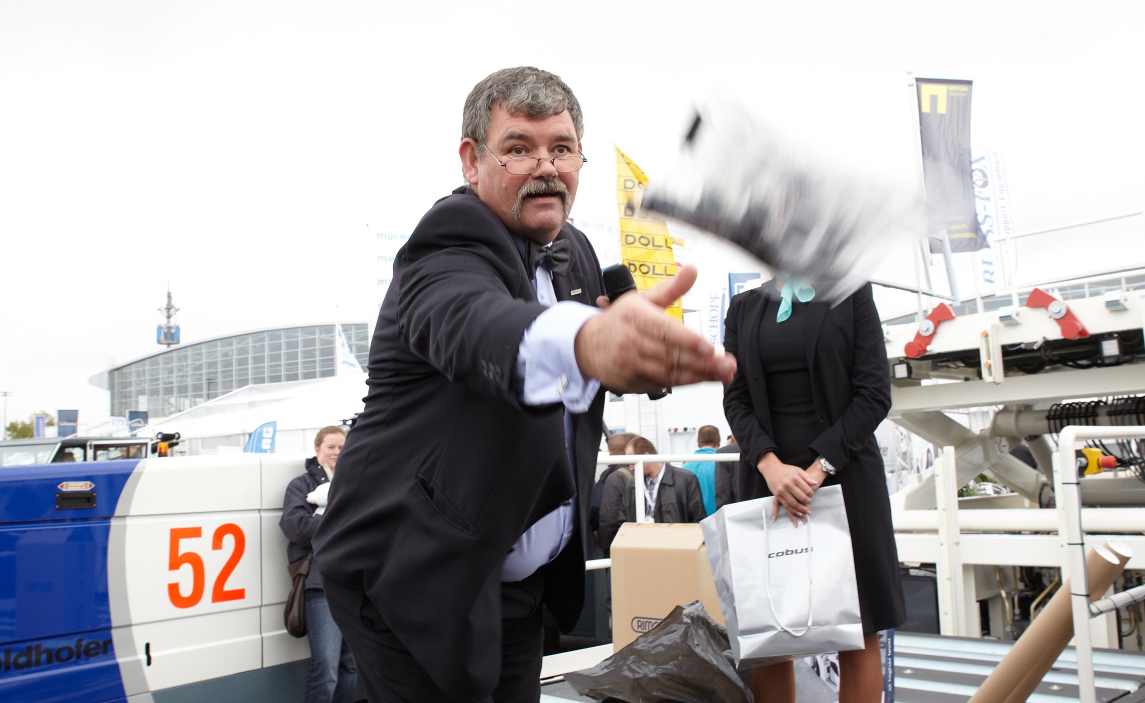 Mann im Anzug wirft ein Give-away in die Menge, im Hintergrund steht eine Hostess vor Flughafenbodengerät