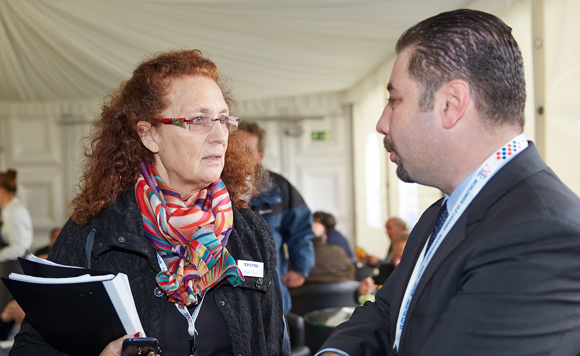Frau mit rotem Lockenhaar und bunter Schal spricht mit einem Mann im Anzug auf einer Messe
