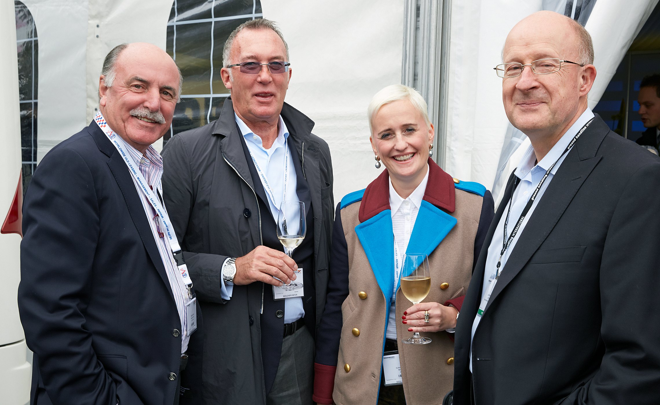 Vier Personen in Businesskleidung stehen vor einem Messestand und halten Weingläser, alle lächeln in die Kamera