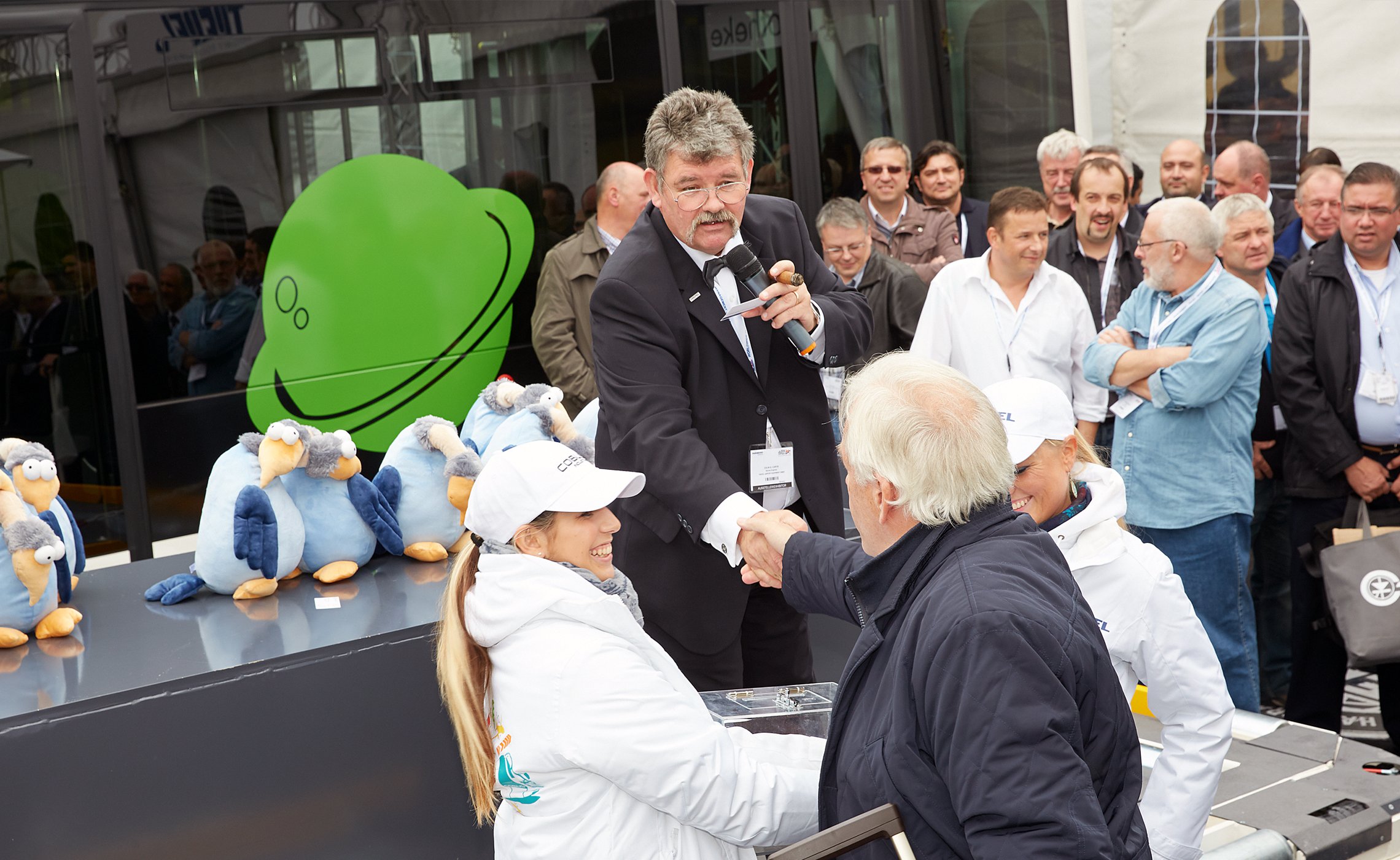 Moderator mit Mikrofon überreicht einem Teilnehmer einen Preis, umgeben von einem Publikum und TREPEL Plüschfiguren