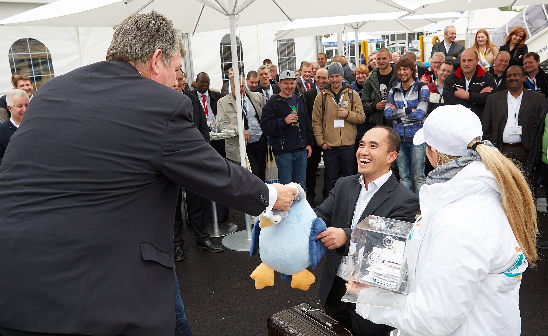 Mann im Anzug erhält eine große Plüschfigur als Gewinn, lachend vor einem applaudierenden Publikum