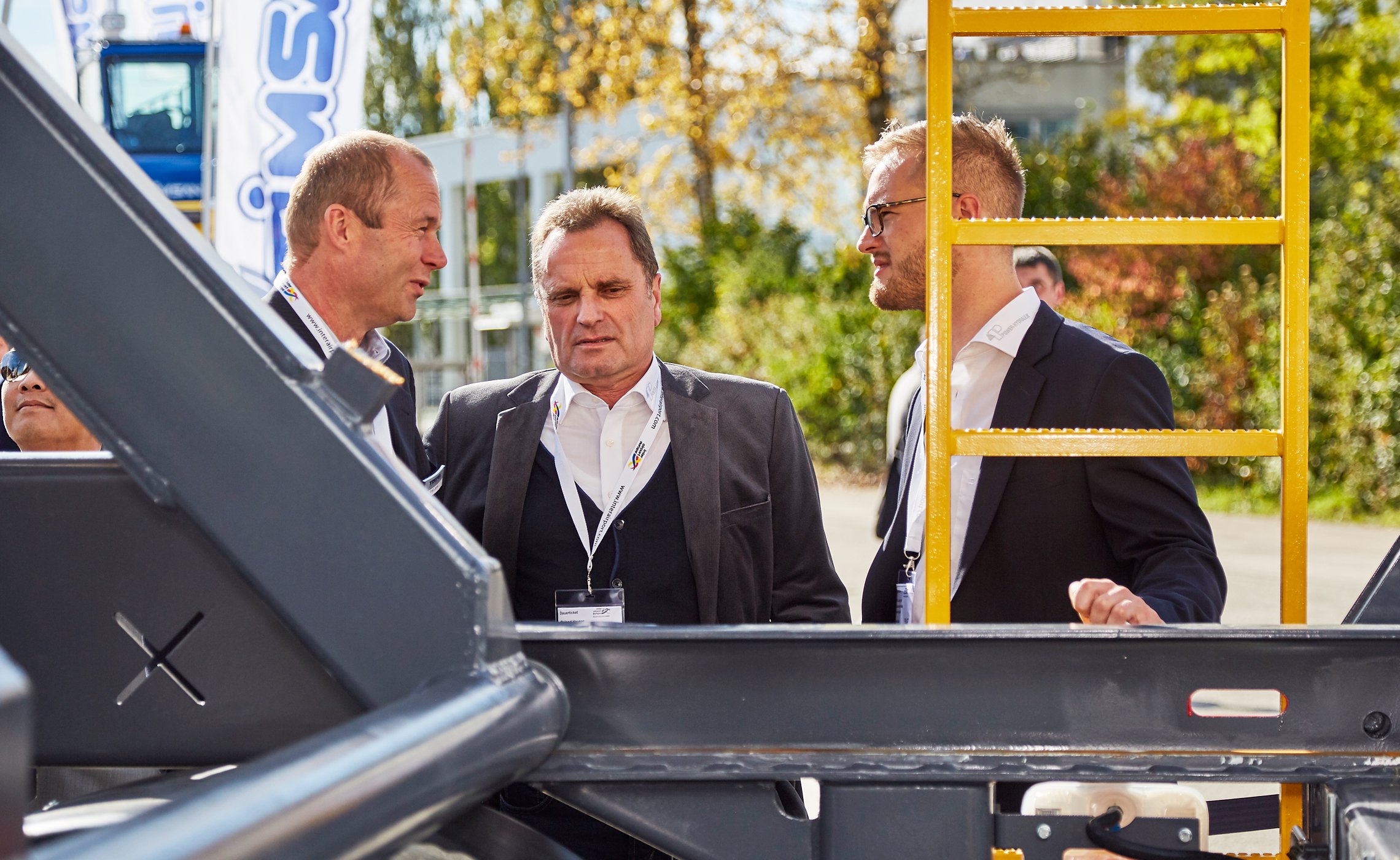 Drei Männer im Anzug unterhalten sich bei Sonnenschein neben einem großen Metallrahmen auf der Inter Airport Europe