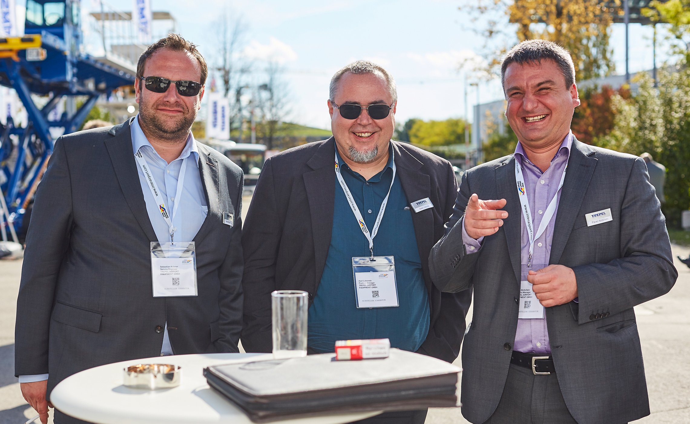 Drei Männer in Anzügen stehen lachend an einem Stehtisch im Freien